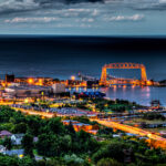 Duluth at night, showcasing the illuminated cityscape, including the Aerial Lift Bridge, with lights reflecting.