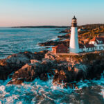 Portland Head Light lighthouse on the coast, with waves crashing against the rocky shoreline and a clear sky above.