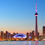 The CN Tower at sunset, illuminated against a vibrant sky with shades of orange & pink, towering over the city skyline.