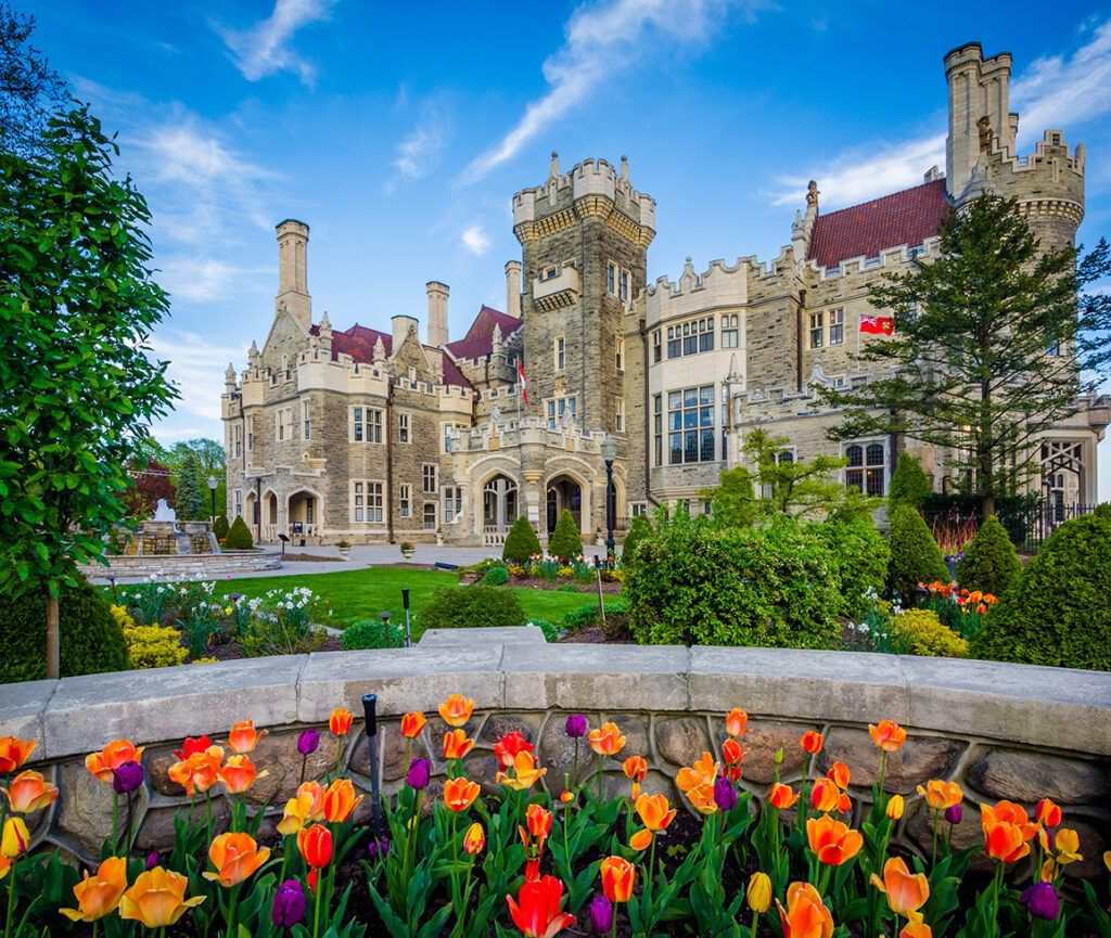 A view of Casa Loma, with its grand architecture and turrets, surrounded by colorful flowers in the front garden.