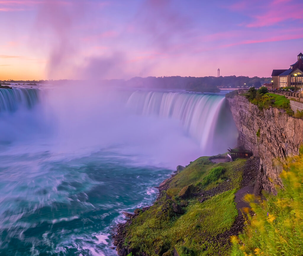 An evening view of Niagara Falls, with the waterfalls illuminated in colorful lights, mist rising.